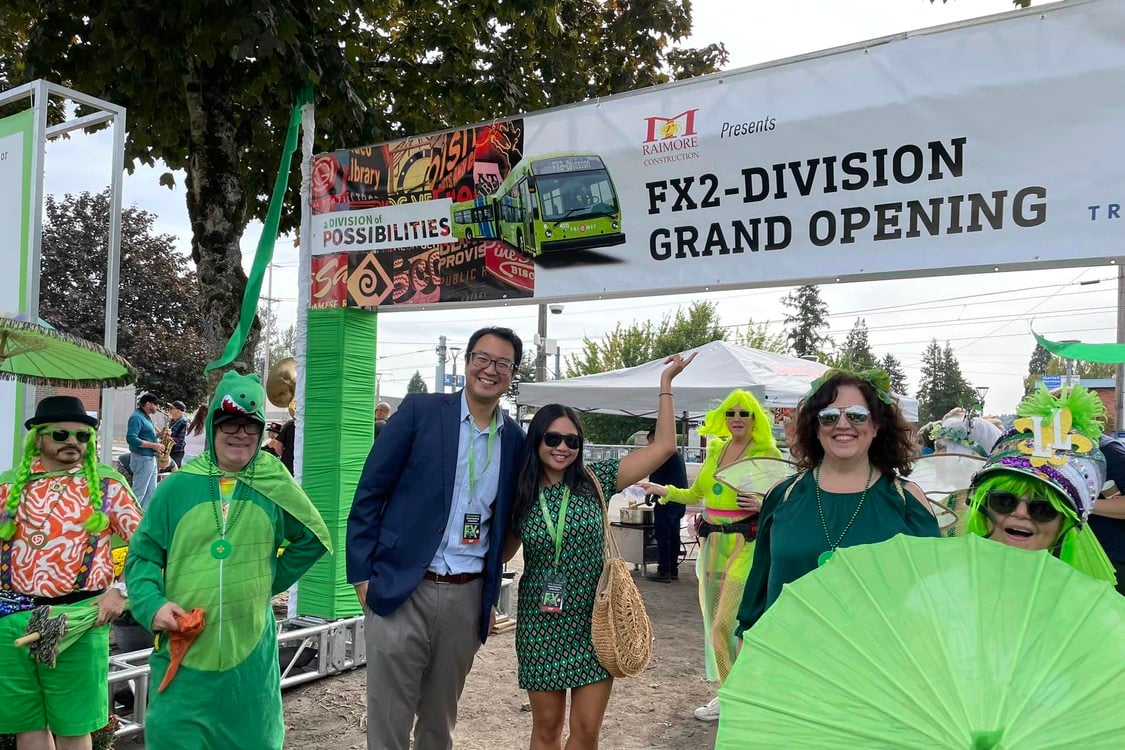 Metro Councilor Duncan Hwang stands next to Kathy Wai under a sign banner displaying "FX2-Division Grand Opening" next to many people dressed in bright green costumes of many styles.