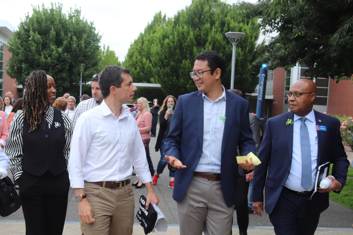 Metro Councilor Duncan Hwang walks alongside Transportation Secretary Pete Buttigieg and other people on a sidewalk at PCC's Southeast campus on 82nd Avenue.