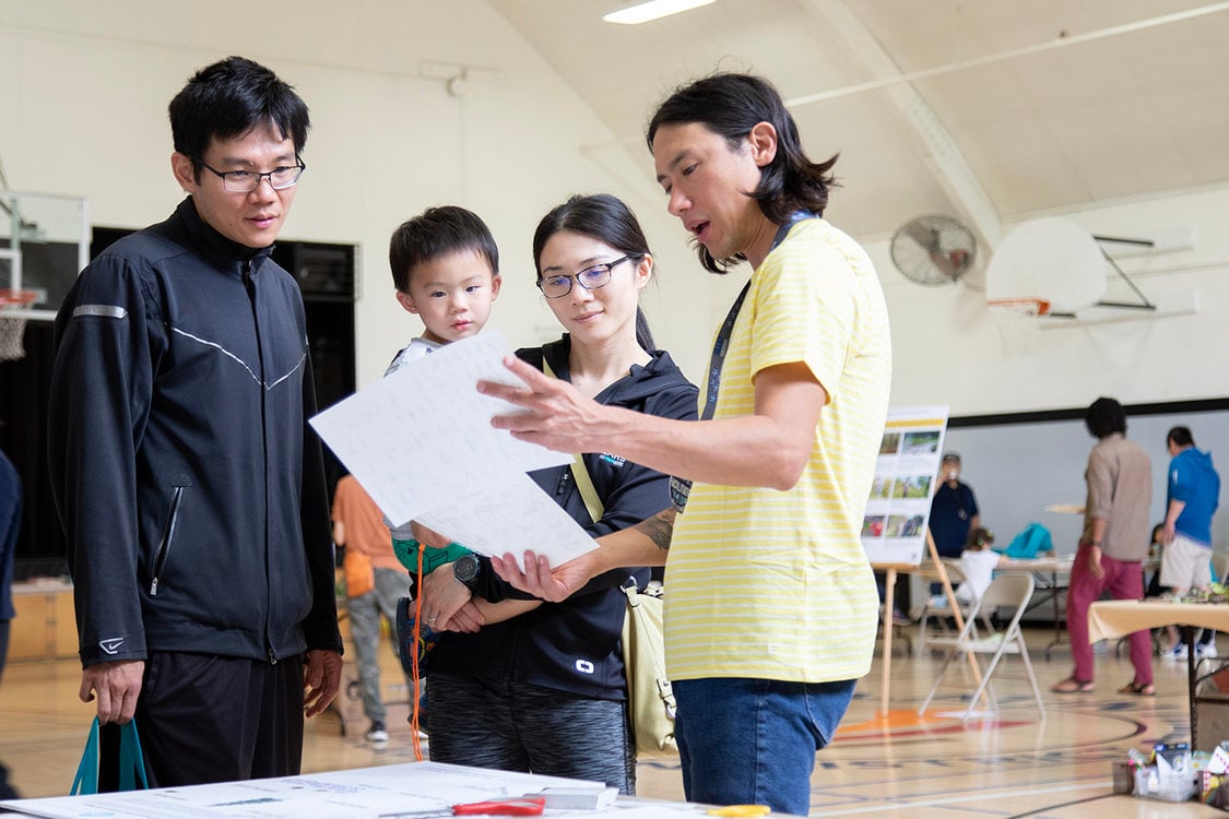 A father, mother and toddler listen to a park designer inside a gym. Other community members are in the background.