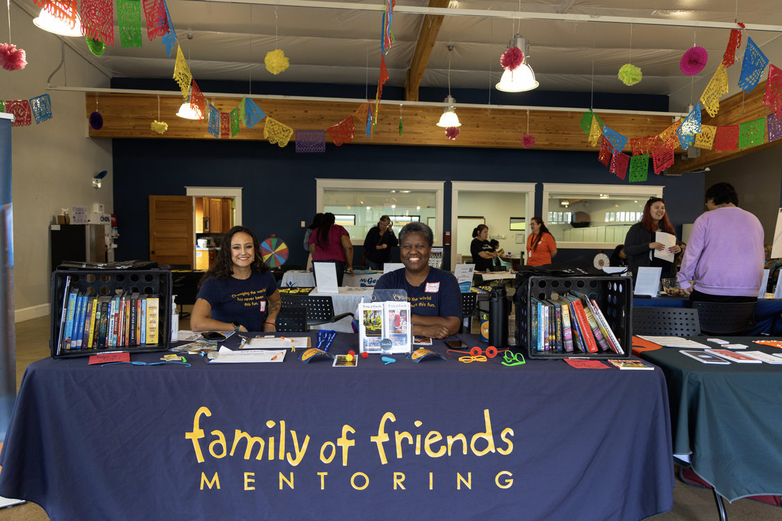 wo people sit in the distance at their table labeled family of friends mentoring at the resource fair. Colorful blue, red, yellow and green banners hang from the ceiling.