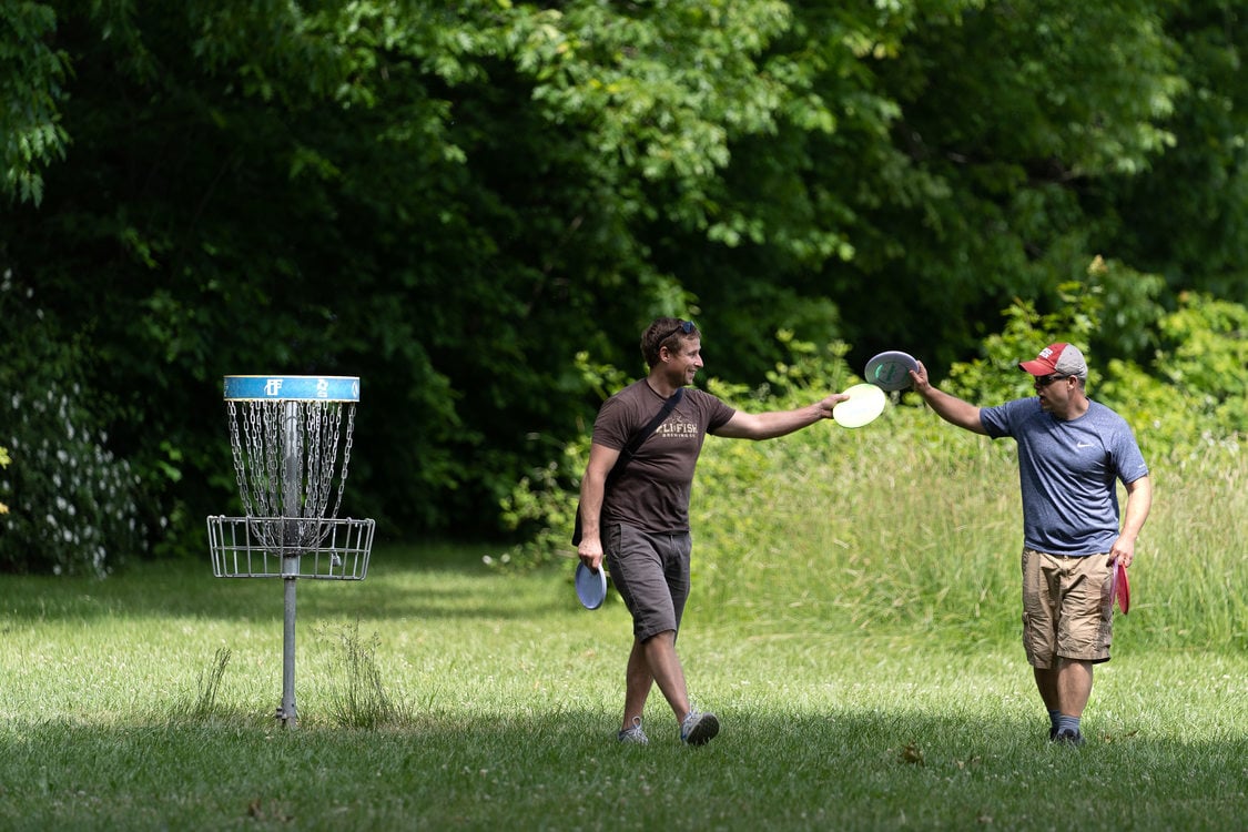 Two men tap discs together while walking near one of the disc baskets at Blue Lake Regional Park disc golf course.