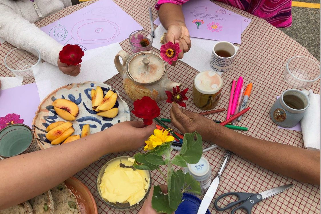 An adult and multiple children show hold their flower creations at the middle of a table full of crafting materials and snacks