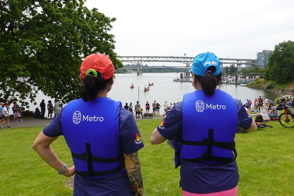 Two women seen from behind wearing Metro life jackets on Poets Beach, watching dragon boats approach on the river