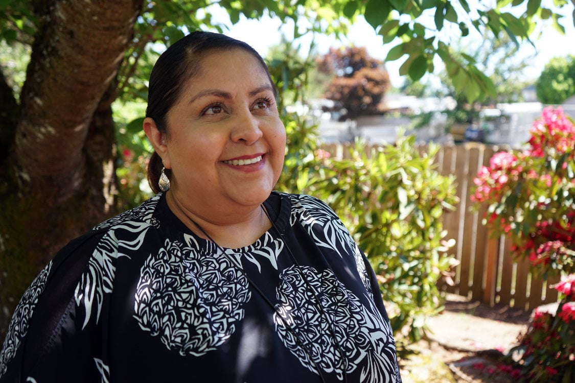 Woman in floral print blouse standing in front of flowering shrubbery and smiling.