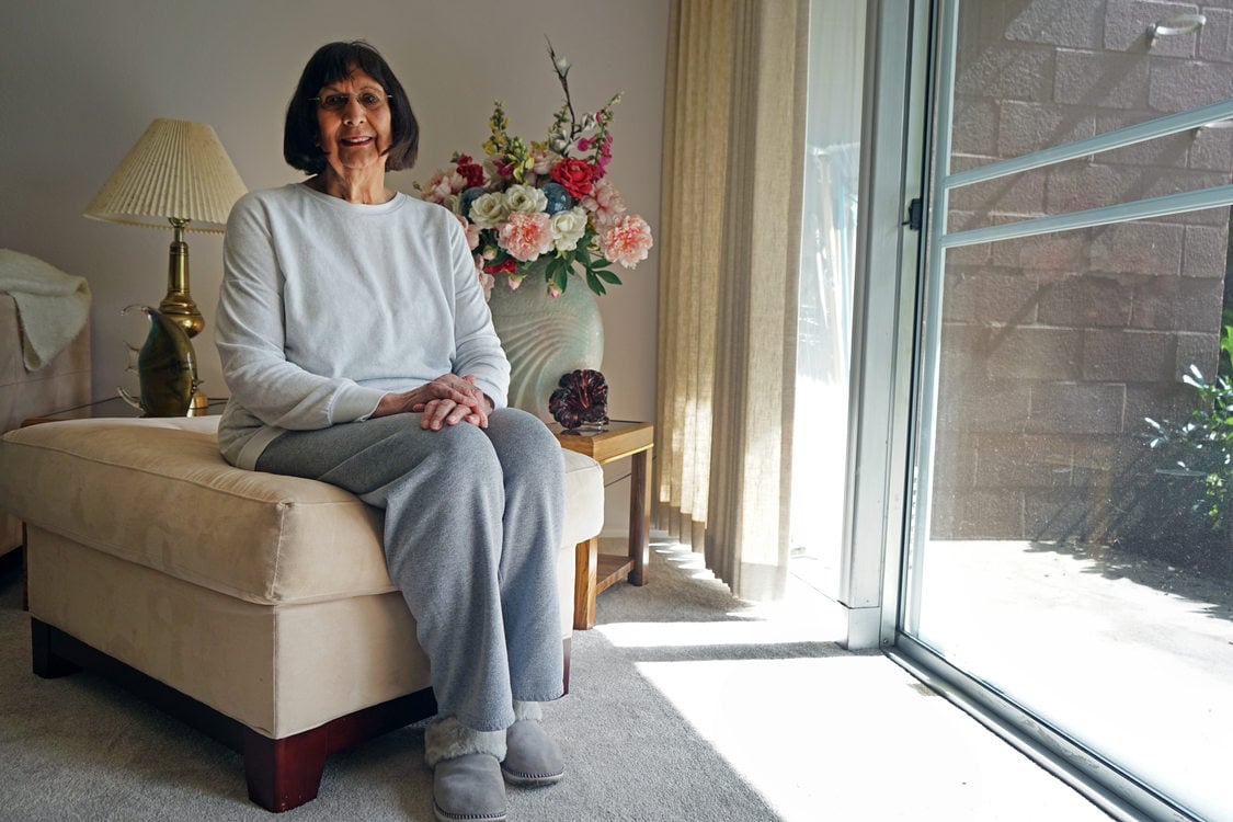 Woman with short dark hair and a white sweatshirt sitting in a livingroom by a glass door.