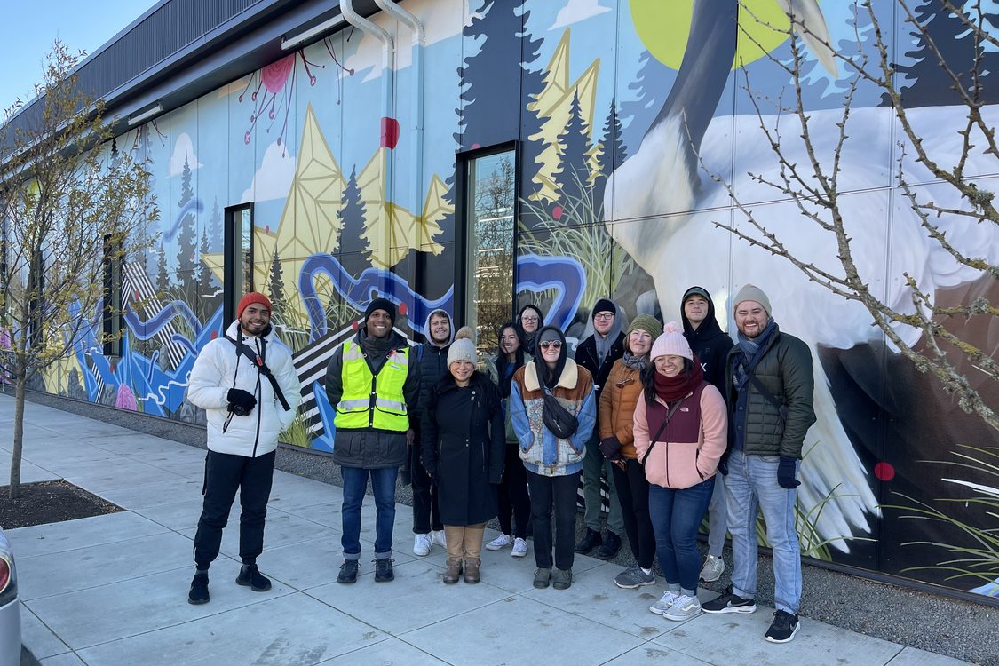 Metro Councilor Ashton Simpson wears a reflective safety vest and poses next to instructor Thuy Tu with a group of students in front of a vibrant wall mural.