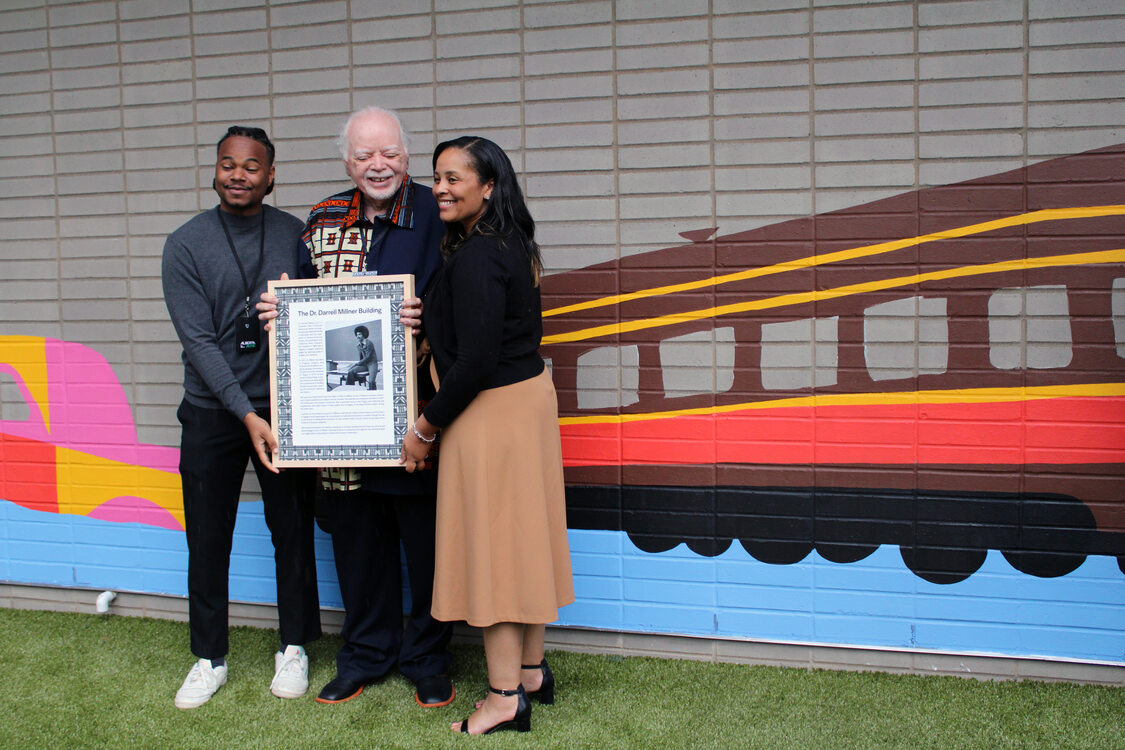 Three people standing in front of a mural featuring a train. The man in the middle holds a framed document and the other two people assist. 