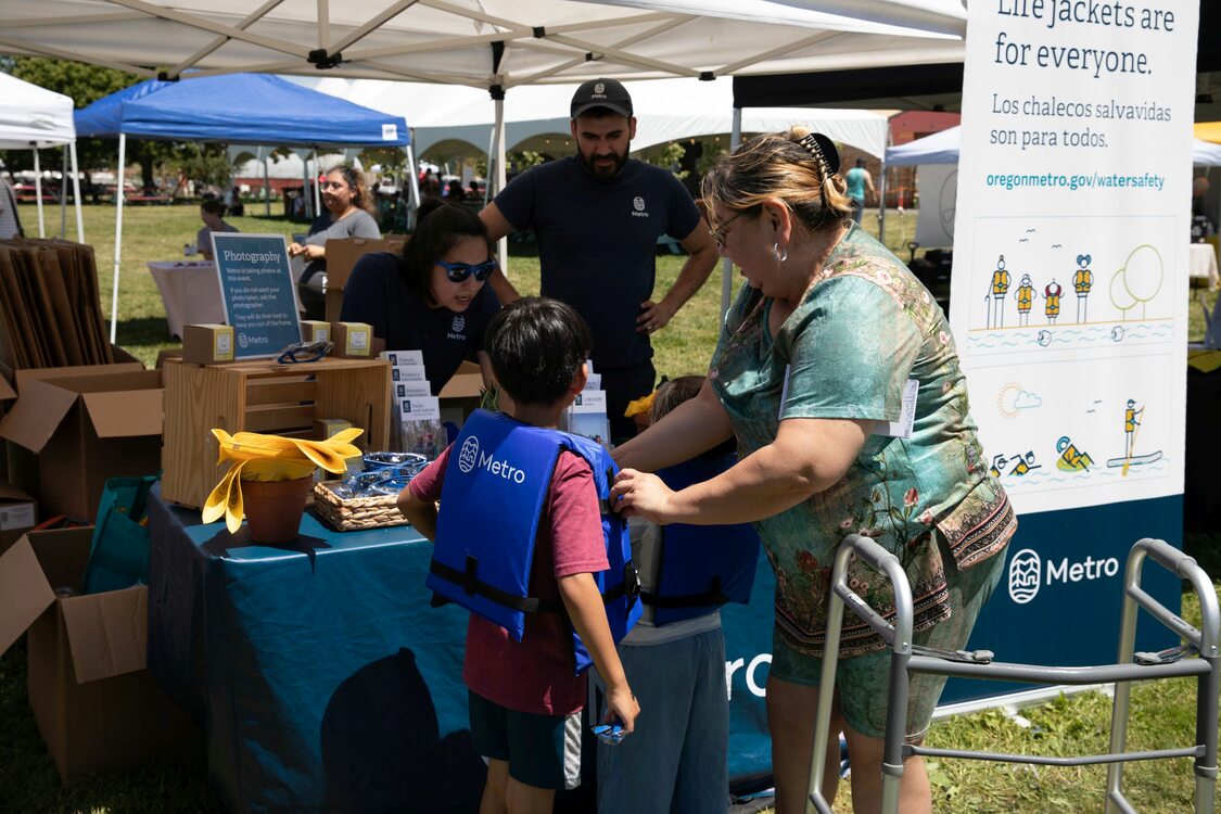 child in a Metro life jacket and an adult stand in front of an outdoor booth staffed by Metro employees, with a banner reading "Life jackets are for everyone" in English and Spanish to one side