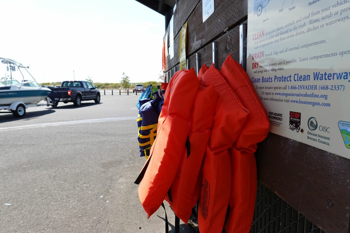 life jackets hanging from a wooden kiosk in a parking lot with a truck pulling a motorboat on a trailer in the background