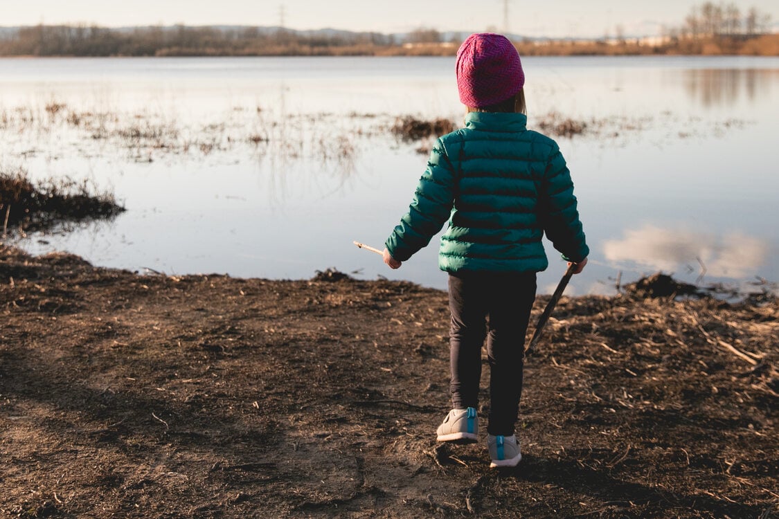 A child wearing a green puffer winter jacket and pink beanie hat stands on the dirt-covered shoreline of a small lake. The sky is sunny with a few puffy clouds in the distance.