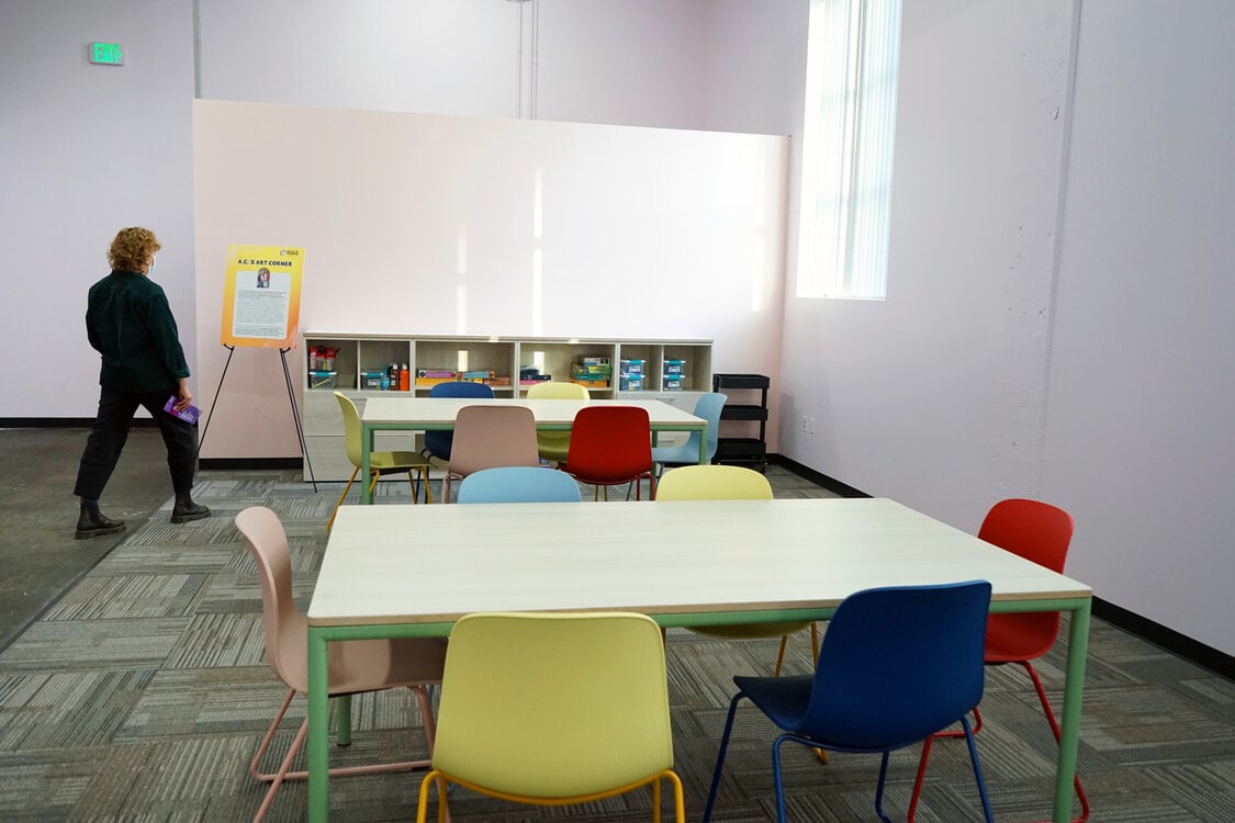 Community art room with colorful chairs. A person in a black jumpsuit reads a sign.