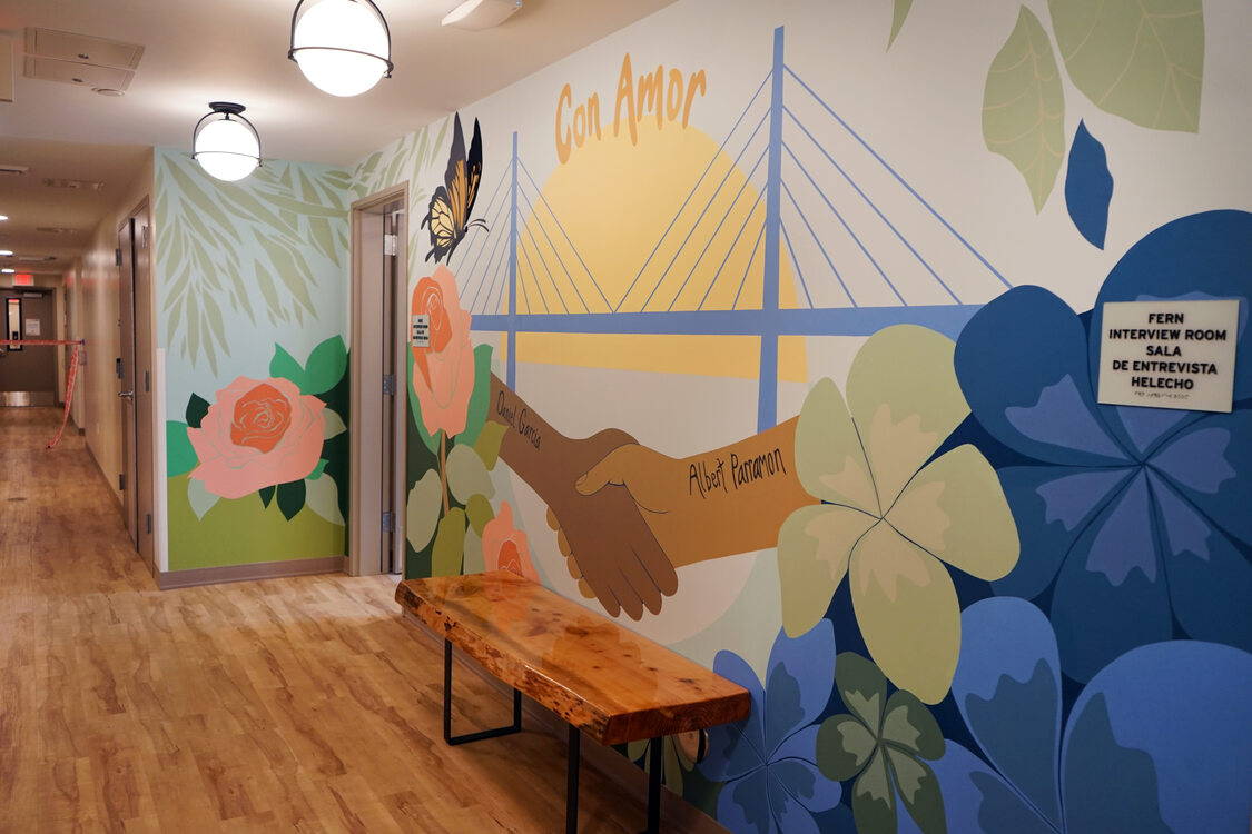 Colorful mural in a hallway featuring a bridge, flowers, a butterfly and clasping hands, with the text "Con Amor." A wooden bench is placed against the wall.