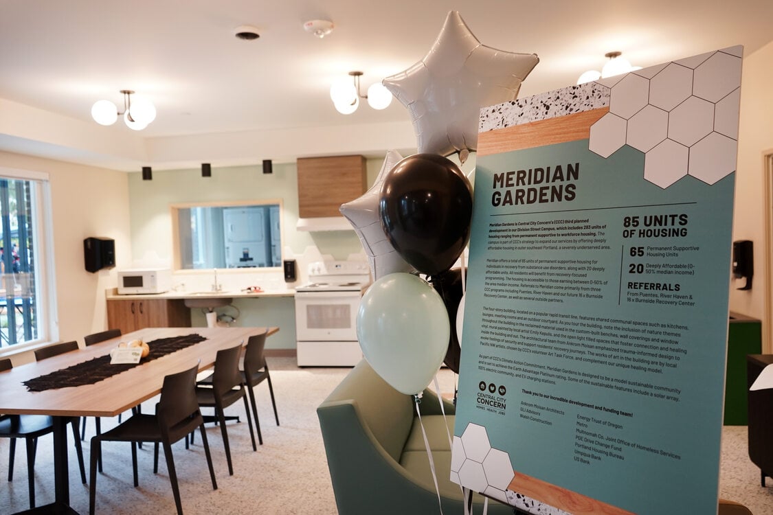 A display board for Meridian Gardens stands inside a kitchen and dining area, with balloons attached. The board details housing units and referral information.