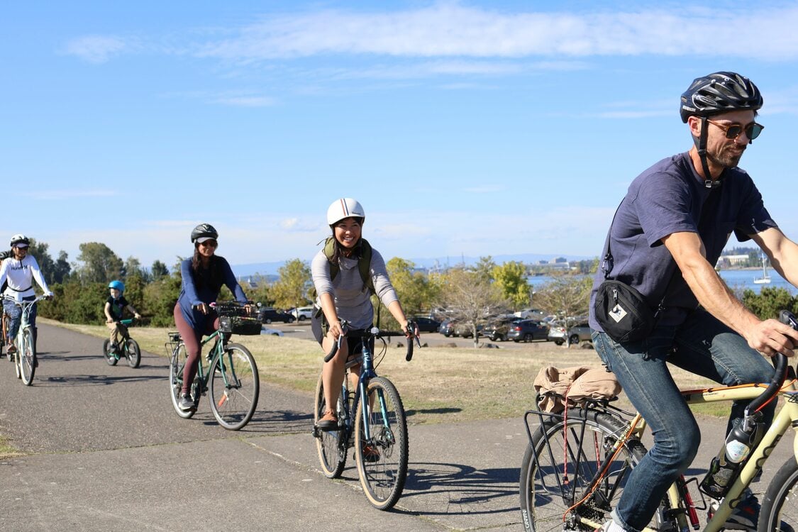 a line of five smiling bicyclists -- one of them a young child -- ride down a bike path on a sunny day, a parking lot and the Columbia River in the background