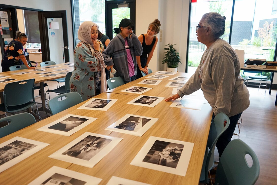 A group of women of various ages gathered around a long table with black and white photo prints spread out on it.