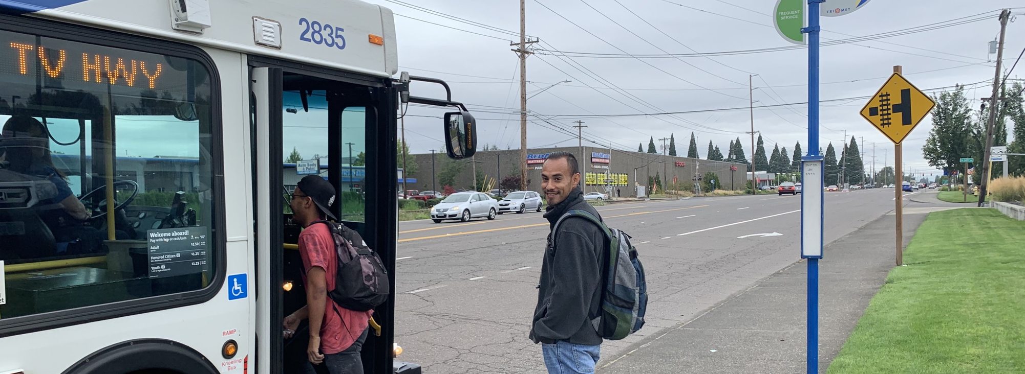 A man wearing a dark grey jacket and backpack stands on a sidewalk, ready to board a TriMet bus on route 57 with the words "TV Highway" in the digital reader board for the bus.