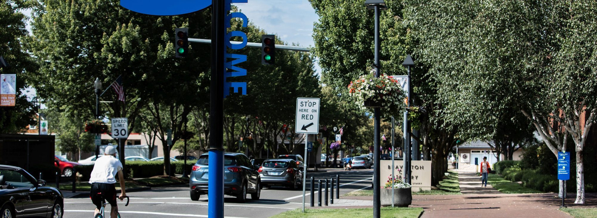 A street, bike lane, sidewalk and welcome sign indicating entry into downtown Beaverton. It is a mostly sunny day, and cars drive on the street. There is a single bike rider in the bike lane. Trees line the road.