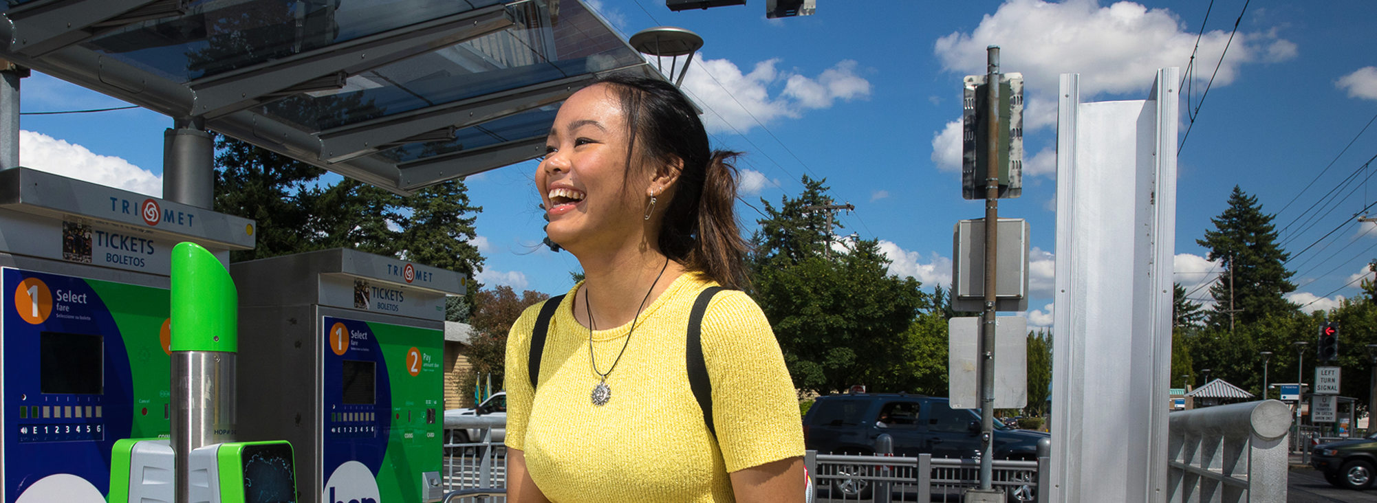 A close up image of a woman in a bright yellow shirt smiling while standing at a TriMet MAX station. 