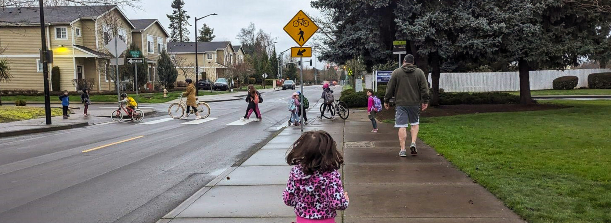 A group of school children and two adults walk or ride their bikes across a crosswalk on a rainy day.