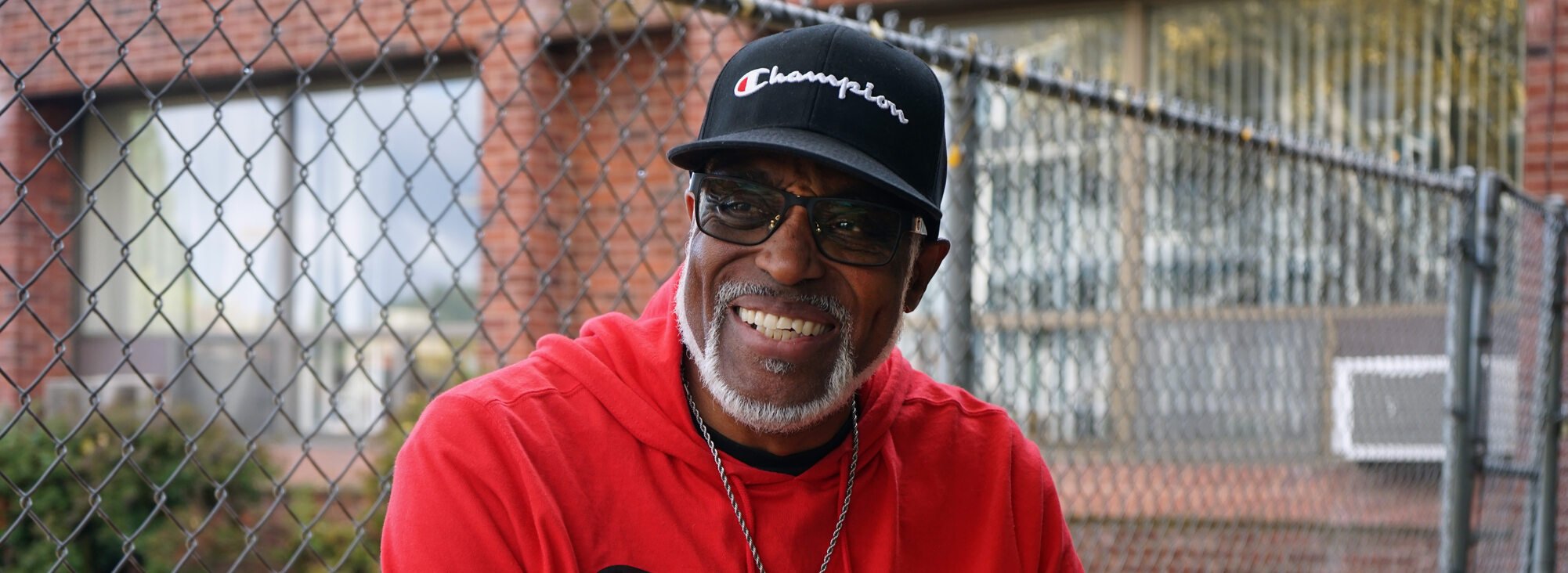 Man in red Champion sweatshirt, black track pants and black ball cap sitting on a bench with fallen gingko leaves and a chain link fence behind him.