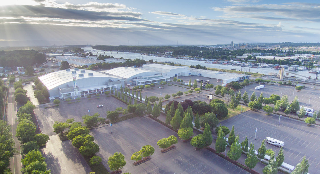 Aerial view of Portland Expo Center, showing the full property, buildings, and parking lots. The view is looking west toward the setting sun, with rays of sunlight streaming through the clouds onto the Expo Center property