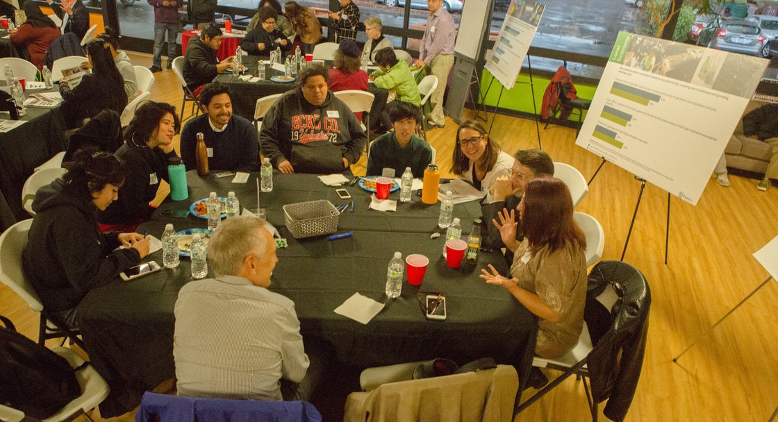 Several groups of people are seated at tables in a meeting room to discuss materials presented on easels placed throughout the room.