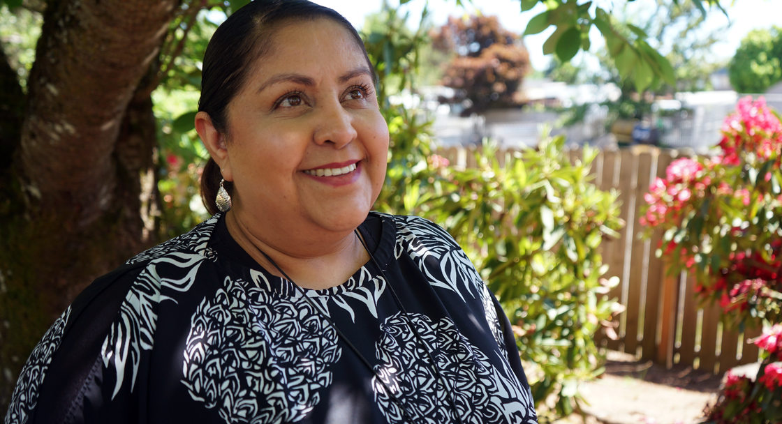 Woman in floral print blouse standing in front of flowering shrubbery and smiling.