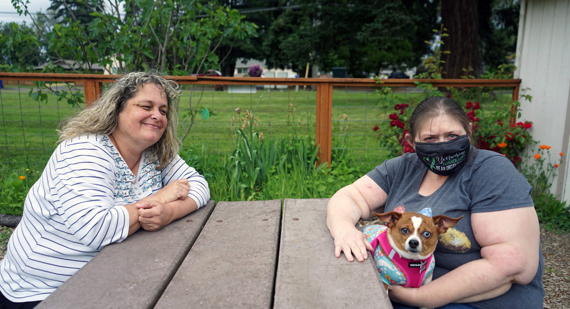 Two women at a picnic table in a grassy setting. One is smiling at the other, who is holding a small dog in her lap.