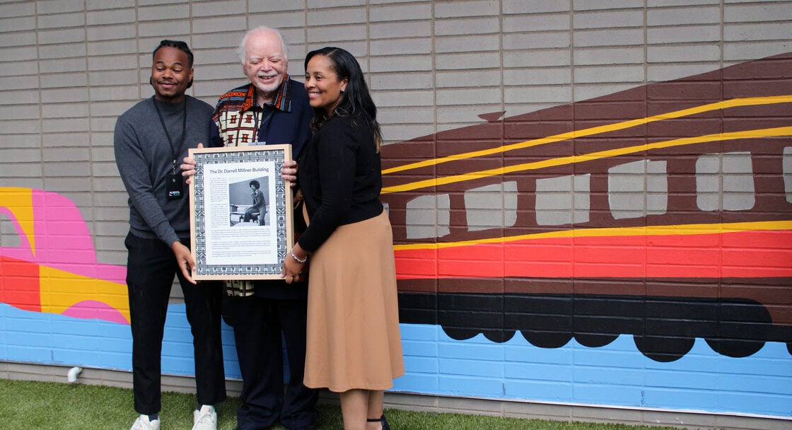 Three people standing in front of a mural featuring a train. The man in the middle holds a framed document and the other two people assist. 