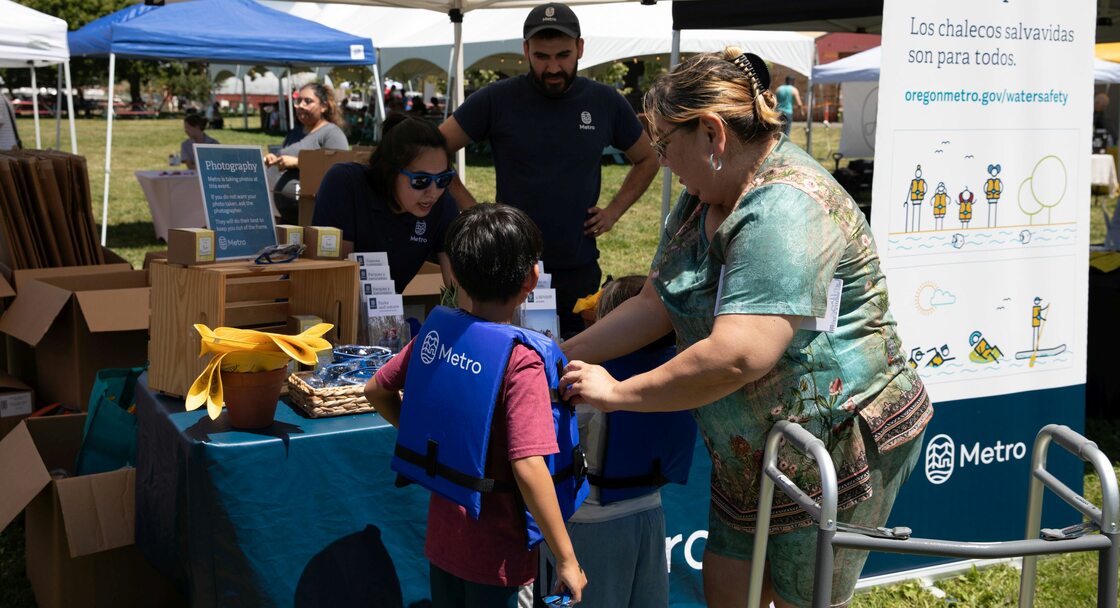 child in a Metro life jacket and an adult stand in front of an outdoor booth staffed by Metro employees, with a banner reading "Life jackets are for everyone" in English and Spanish to one side