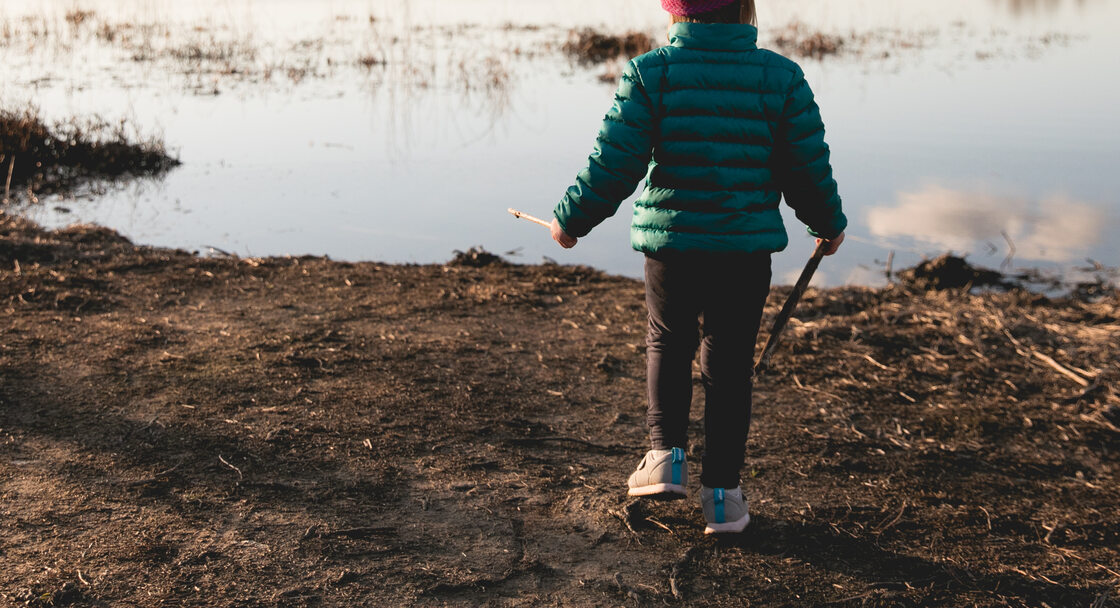 A child wearing a green puffer winter jacket and pink beanie hat stands on the dirt-covered shoreline of a small lake. The sky is sunny with a few puffy clouds in the distance.